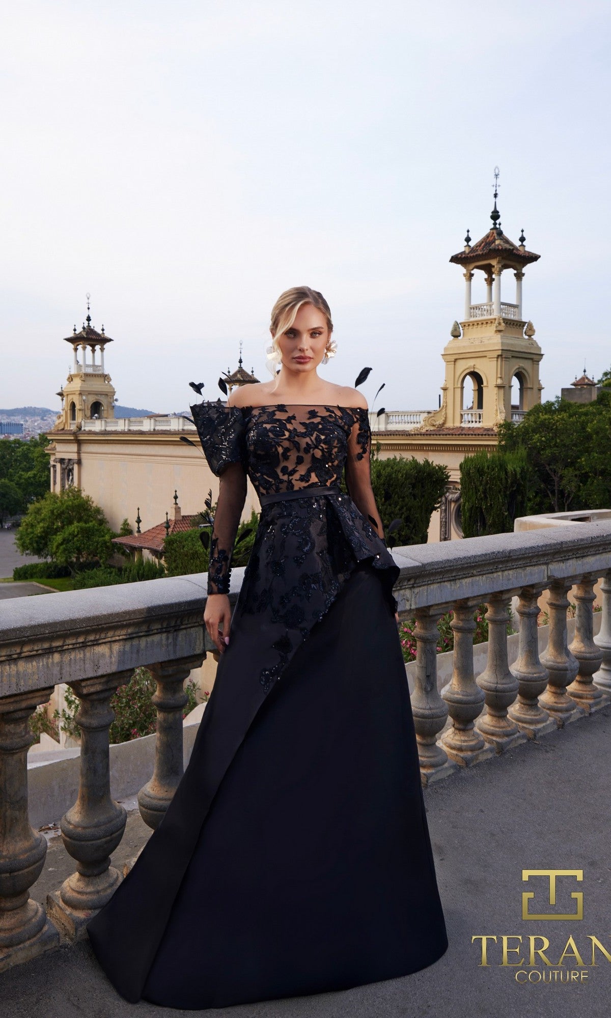 A woman wears the Long Formal Dress: Terani 261M6410 in Black Nude, featuring elegant sheer floral detailing, as she stands on a stone terrace with ornate buildings and trees behind her. The Terani Couture logo highlights this mother of the bride gown.
